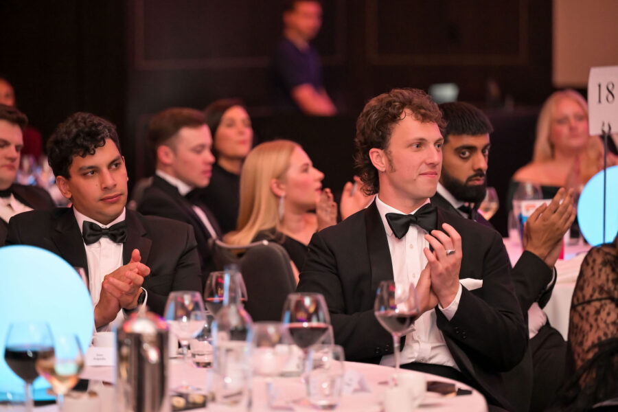 “Attendees in black-tie attire sit at round banquet tables and applaud during a formal awards event, with wine glasses, place settings, and table numbers visible.”