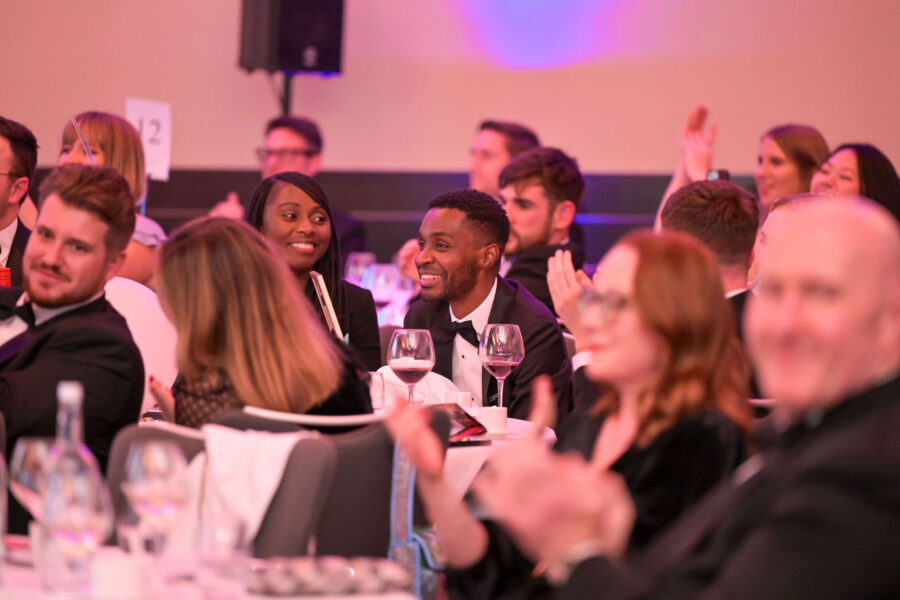 “Attendees at a formal awards event sit at a round table and applaud, with wine glasses, bottles, and tableware visible under pink ambient lighting.”
