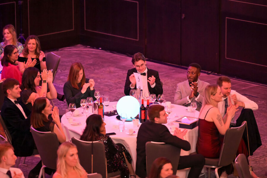 “Attendees seated at a round banquet table applaud during a formal awards event, with wine glasses, bottles, and a table number visible, illuminated by pink ambient lighting.”