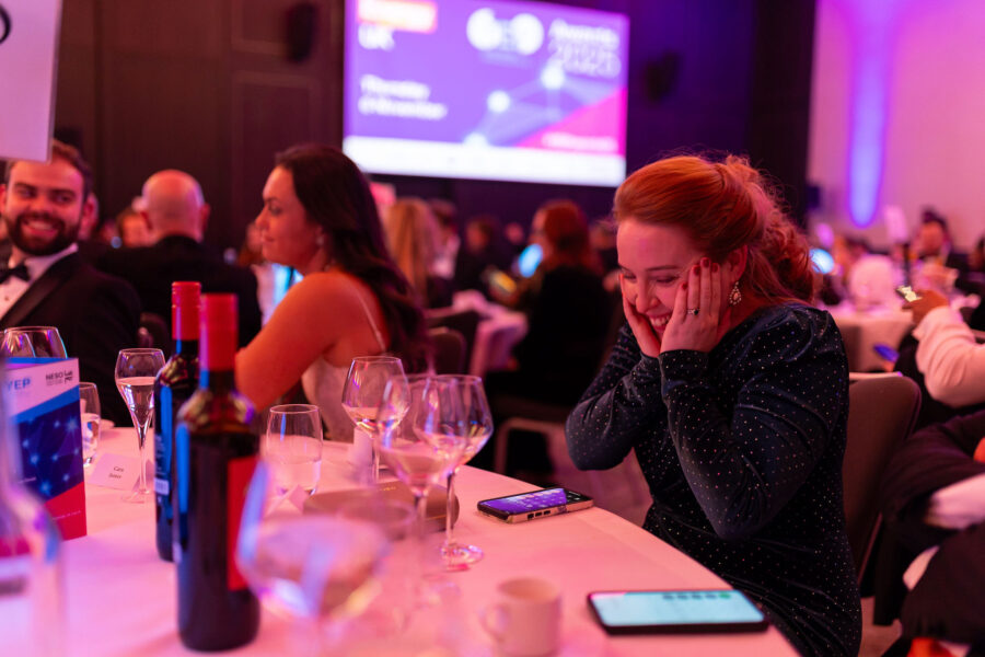 “Guests sit at a round banquet table with wine glasses, a bottle, and table materials visible, while one attendee rests hands against face during a formal awards event with purple stage lighting in the background.”