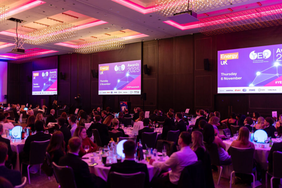 A large audience seated at round banquet tables during a formal awards ceremony, with purple and pink lighting and multiple screens displaying ‘Energy UK YEP Awards 2025’ and event details