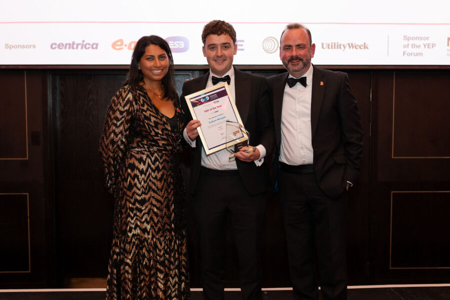 Three attendees dressed in formal evening wear stand together on stage, with the centre person holding a framed certificate and glass award. A screen behind displays ‘Energy UK YEP Awards 2025’ and sponsor logos.